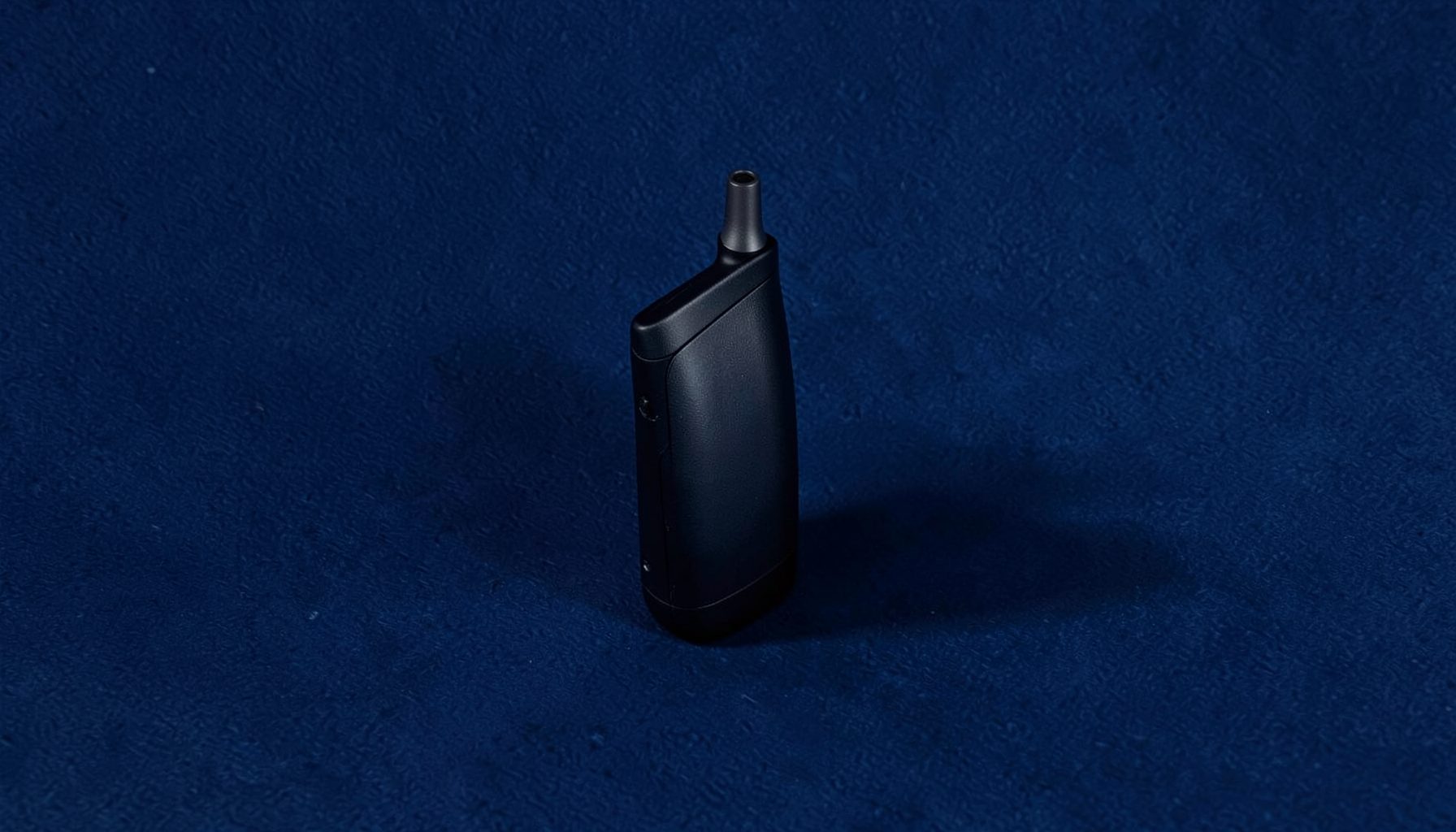 A textured navy-blue background with a single vape device casting a soft shadow, lit from the side to emphasize its sleek profile
